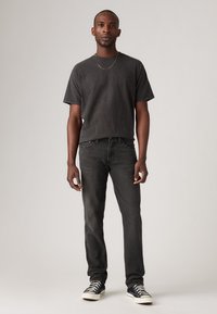 Svart kortermet t-skjorte med avslappet passform, kombinert med mørke smale jeans og svarte og hvite sneakers mot en ensfarget bakgrunn.