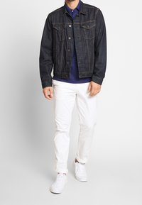Veste en denim foncé avec deux poches poitrine, coutures orange, portée sur une chemise bleu marine. Assortie avec un pantalon blanc retroussé et des baskets blanches.