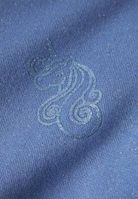 Mėlynas audinys su subtiliu, blizgančiu vienaragio logotipu sukimo dizainu, švelnios tekstūros ir smulkios mezgimo rašto visame paviršiuje.