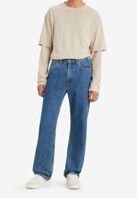 Beige T-shirt med kort ärm i lager över en skjorta med lång ärm, tillsammans med vida blå denimjeans och vita sneakers.