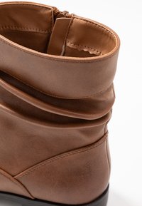 Bottine en cuir marron avec un design froncé, texture lisse et semelle en caoutchouc noir ; elle possède une fermeture éclair et des coutures visibles.