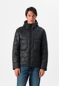 Casaco acolchoado preto com capuz, apresentando dois bolsos frontais, fecho éclair na frente e uma textura suave e brilhante. Usado sobre um par de jeans de ganga.