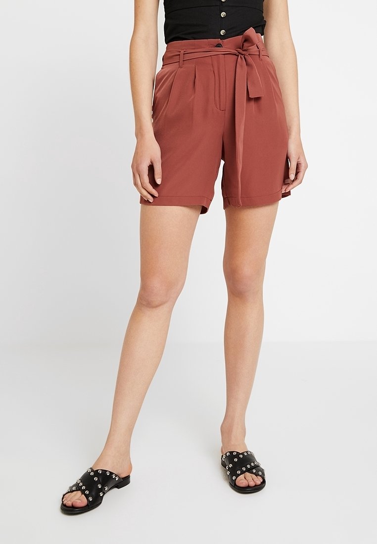 Femme portant un short taille haute couleur rouille avec une ceinture nouée et des sandales noires cloutées, debout devant un fond uni.