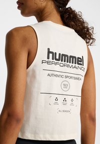 White cropped tank top made of cotton, featuring bold black text on the back: "hummel PERFORMANCE" and various symbols regarding functionality.