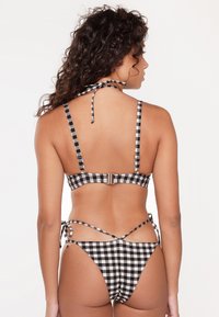 Le bikini en vichy noir et blanc présente un haut à col halter, des liens arrière ajustables et une fermeture à cliquet sur le haut, soulignant un design ajusté.