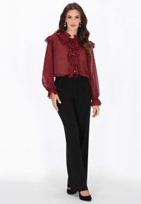 Bordó áttetsző blúz fodros díszítéssel, gombos részletekkel és hosszú ujjakkal, fekete szabott nadrággal és fekete magassarkúval párosítva.