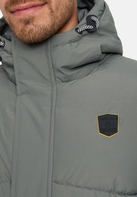 Gros plan sur le bas du visage et le cou d'un homme portant une veste matelassée grise avec capuche et un patch logo noir et jaune d'Indicode.