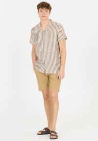 Kortärmad skjorta med knappar i vertikala ränder i beige och brunt, tillsammans med sandfärgade shorts. Modellen har på sig bruna sandaler.