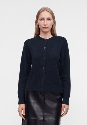 Femme aux cheveux blonds et raides portant un pull bleu foncé boutonné et une jupe en cuir noire sur un fond clair uni.