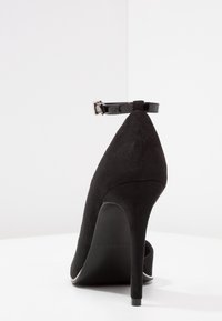Scarpa nera in suede con tacco alto, punta appuntita e cinturino alla caviglia regolabile, caratterizzata da un design elegante e una texture liscia.