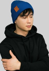 Niebieska, prążkowana czapka z daszkiem z skórzaną naszywką, noszona z czarnym, kapturkiem swetrem. Materiał jest teksturowany, co zapewnia dopasowanie.