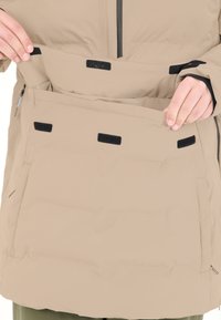 Beigefärgad isolerad jacka med en bred framficka, svarta kardborrestängningar och en smidig textur. Har en zip-ficka på sidan.