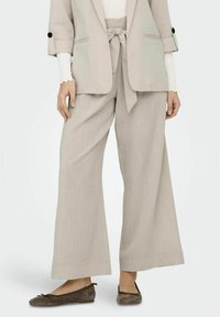 Pantalons larges en mélange de lin beige clair avec une ceinture en tissu, finition texturée, associés à des ballerines en daim marron foncé.