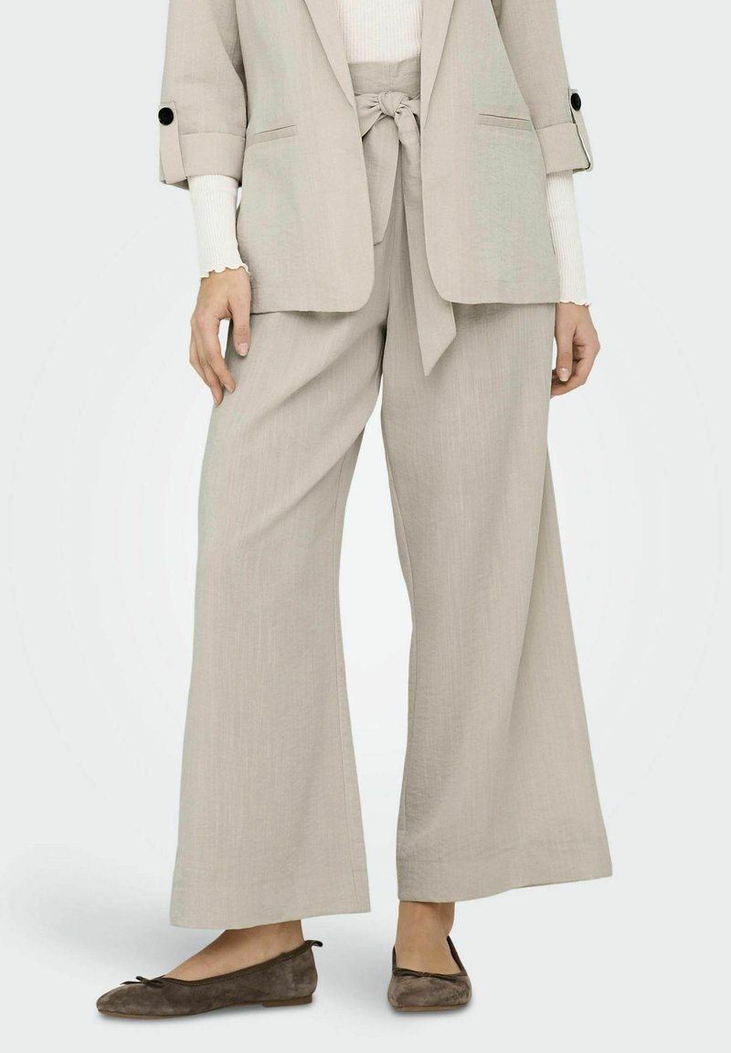Pantalons larges en mélange de lin beige clair avec une ceinture en tissu, finition texturée, associés à des ballerines en daim marron foncé.