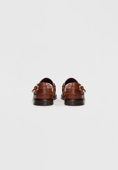 Kastanjebrown läderloafers med guldspännen visade från baksidan på en enkel ljusgrå bakgrund.