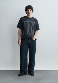 Uomo con capelli ricci, indossa una t-shirt nera da tour con grafica, jeans scuri larghi e scarpe nere, in piedi contro uno sfondo grigio uniforme.