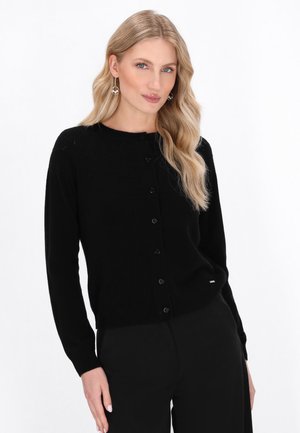 Schwarze gestrickte Strickjacke mit rundem Ausschnitt, langen Ärmeln und Knopfverschluss. Glatte Textur mit einem subtilen Muster, kombiniert mit schwarzen Hosen.