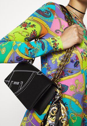 Černá obdélníková taška s bílými sešitými detaily a štítkem "Versace Jeans Couture", držená zlatým řetízkem; viditelný barevně vzorovaný rukáv.