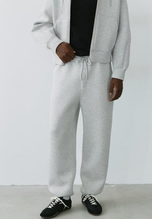 Person iført lysegrå joggingbukser med snøre, matchende zip-up hættetrøje, sort trøje og sorte sneakers med hvide snørebånd mod en ensfarvet baggrund.