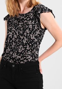 Svart topp med korta, vågiga ärmar som har ett blommönster i rosa och vitt. Lätt material, rund halsringning och en avslappnad passform.