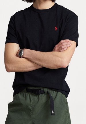 Mann mit schwarzem Kurzarmhemd mit rotem Logo, grüner Hose und schwarzem Gürtel mit Logo sowie einer Armbanduhr, die Arme gekreuzt.