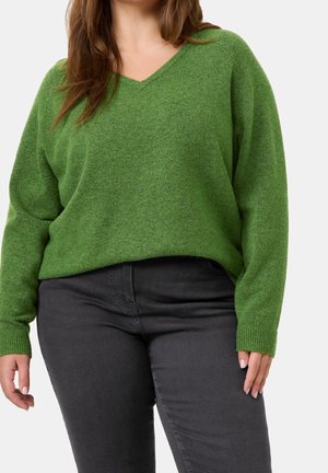 Femme portant un pull vert ample à col en V et un pantalon gris foncé ajusté, les mains détendues le long du corps.
