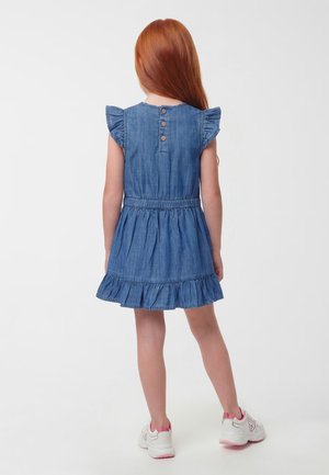 Fille aux longs cheveux roux portant une robe en jean bleue à volants et des baskets blanches, debout de dos sur un fond uni.