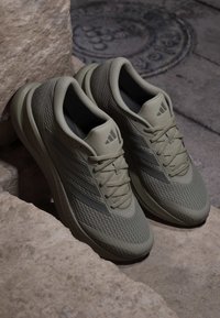 Ζευγάρι ανοιχτόχρωμα γκρι παπούτσια Adidas για τρέξιμο με διχτυωτό ύφασμα και κορδόνια τοποθετημένα σε πέτρινα σκαλοπάτια κοντά σε κάλυμμα φρεατίου.