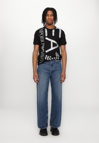 Tricou negru cu imprimeu mare alb, purtat cu blugi albaștri cu croială lejeră. Blugii au o spălare deschisă și o textură ușor decolorată. Pantofi negri.
