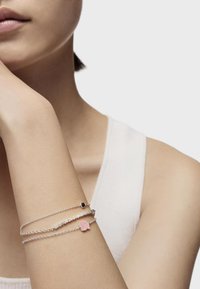 Bracciale in argento a tre fili, con un charm geometrico rosa e una piccola perla nera; texture liscia sulla pelle nuda.