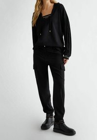 Schwarzer Hoodie mit strukturierten Ärmeln, V-Ausschnitt und Kordelzügen; kombiniert mit schwarzen Cargo-Hosen mit Seitentaschen; schwarze Schuhe mit Akzenten.