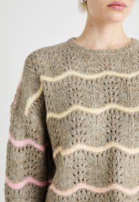 Brede gebreide trui in beige met golfpatroon; heeft pastelroze en gele strepen. Gestructureerd met een losse weefstructuur, ronde halslijn en verlaagde schouders.