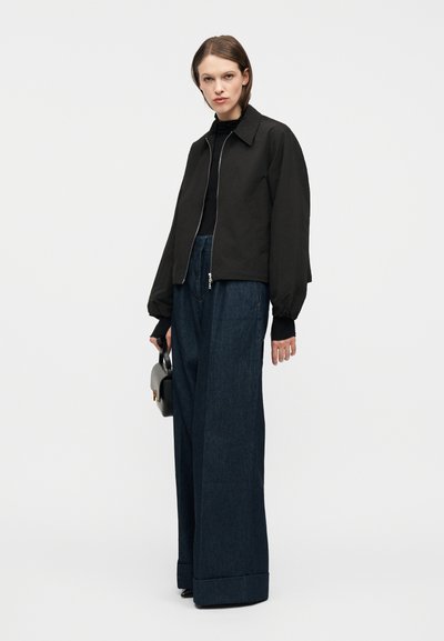 Γυναίκα που φοράει μαύρο μπουφάν με φερμουάρ, μαύρο τοπ, μπλε σκούρο φαρδιά τζιν, κρατώντας μια μικρή τσάντα και στέκεται μπροστά σε λευκό φόντο.