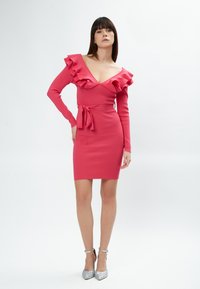 Abito aderente rosa con maniche lunghe, scollatura a V profonda, spalle con volant e cintura annodata in vita. Indossato con tacchi argentati glitterati.