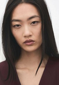 Zbliżenie portretu osoby o długich, ciemnych włosach i neutralnym makijażu, ubranej w głęboko bordową bluzkę. Charakterystyczne cechy to gładka skóra i subtelne pieprzyki.