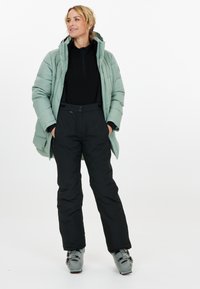 Puffig mintgrön jacka med huva, svart zip-up tröja och svarta skidbyxor. Grå skidstövlar med spännen. Avslappnad pose mot en vit bakgrund.