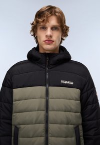 Giacca imbottita nera e verde oliva con chiusura a zip, cappuccio e dettaglio del logo sul petto. Presenta una texture trapuntata e tasche laterali.