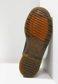 Semelle de chaussure en cuir noir avec une bande de roulement en caoutchouc translucide marron, montrant des grips rectangulaires et des détails de marque en relief.