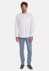 Witte linnen blouse met lange mouwen en een voorzak, gecombineerd met lichtblauwe slim-fit broek en donkere sneakers met witte zolen.