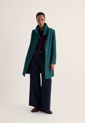 Femme debout portant un manteau boutonné bleu sarcelle, un pantalon large foncé, un haut violet et des chaussures marron sur un fond beige uni.