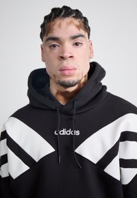Svart hoodie med vita chevronmönster på axlarna, en kapuschong med dragsko och "adidas"-logotyp på bröstet. Mjuk textur.