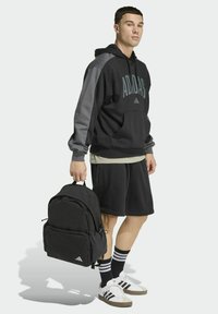 Sudadera con capucha negra de Adidas con mangas grises, pantalones cortos negros, mochila negra y calcetines con rayas blancas, combinados con zapatillas negras y blancas.