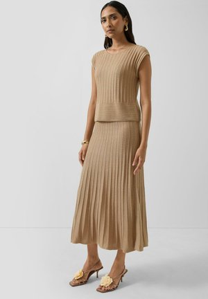 Kobieta ubrana w bezrękawnik w beżowym prążkowanym dzianym topie oraz dopasowaną plisowaną spódnicę midi, do tego brązowe sandały na obcasie z dużymi kwiatowymi ozdobami.