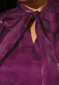 Blusa di satin viola con un grande fiocco al collo, apertura a chiave e una texture liscia e lucida con una leggera lucentezza.