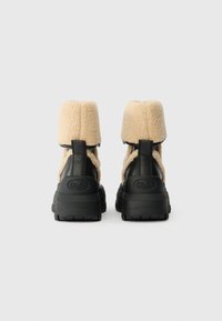 Μαύρες και μπεζ μποτες μέχρι τον αστράγαλο με επάνω μέρος από αφρώδη fleece, καουτσούκ σόλα και χαρακτηριστικές λεπτομέρειες λογότυπου στη φτέρνα. Όγκος και υφή σχεδίου.