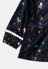 Svart regnjakke med fargerike mønstre av enhjørninger og sløyfer, elastiske mansjetter og en reflekterende hvit stripe langs kanten. Glatt, glansfullt stoff.