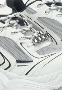 Witte en grijze sporty schoenen met een bovenwerk van mesh, zwarte accenten en decoratieve zilveren hardware over de veters. Gestructureerde zool.
