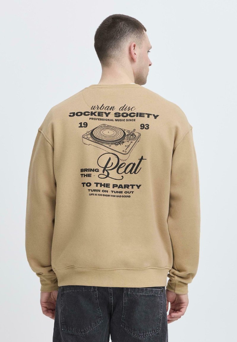 Uomo che indossa un felpa beige con grafica nera e testo sulla musica e un giradischi sul retro, abbinata a jeans scuri.