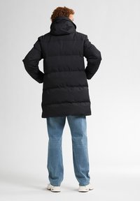 Zwarte lange pufferjas met capuchon, voorzien van een gewatteerd ontwerp en elastische cuffs, gecombineerd met lichtblauwe straight-leg jeans en witte sneakers.