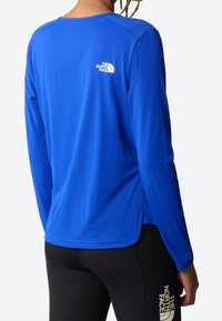 Osoba v modrém tričku s dlouhými rukávy The North Face a černých legínách s logem The North Face na stehně, zobrazená zezadu.